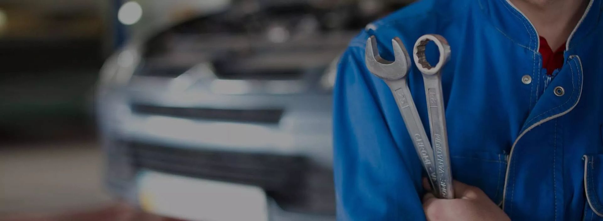 Kfz-Mechaniker in blauem Overall hält zwei Schraubenschlüssel vor einem geöffneten Fahrzeug in der Werkstatt